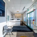Modern dental office reception hallway: white walls, glass doors, waiting chairs, mounted TV, and front desk visible
