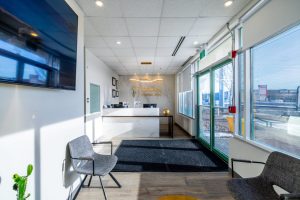 Modern dental office reception hallway: white walls, glass doors, waiting chairs, mounted TV, and front desk visible